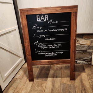 May include: A wooden A-frame chalkboard sign with a black chalkboard surface. The sign has the word "BAR" written in white letters at the top and a list of drinks written in white letters below. The drinks include beer, liquor, mixers, and wine.