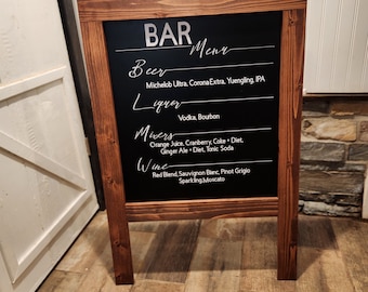 May include: A wooden A-frame chalkboard sign with a black chalkboard surface. The sign has the word "BAR" written in white letters at the top and a list of drinks written in white letters below. The drinks include beer, liquor, mixers, and wine.