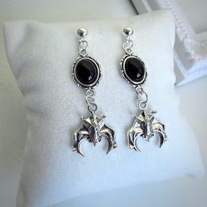 Könnte beinhalten: Ein Paar silberfarbene Ohrhänger mit schwarzen, ovalen Steinen und Fledermaus-Anhängern. Die Ohrringe haben ein Stecker- und Ketten-Design mit einem kleinen Ball oben. Die Fledermaus-Anhänger sind detailliert mit Flügeln und Gesichtern.
