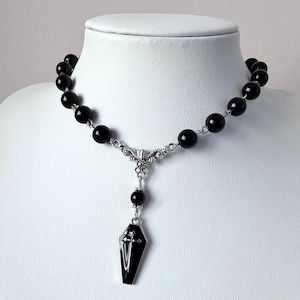 Può includere: Una collana di perline nere con un ciondolo argentato. Il ciondolo è a forma di bara nera con una croce argentata. La collana è esposta su un busto di manichino bianco.