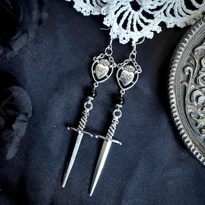 May include: Silver-toned dangle earrings featuring a heart and crown design, with black beads and dagger pendants. The earrings are displayed on a dark background with a lace trim and a decorative silver plate.