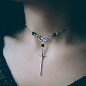 Può includere: Collana color argento con un ciondolo a forma di spada. La collana ha una catena delicata con accenti di perline nere e un elemento centrale ornato. Il ciondolo a spada è dettagliato con un'impugnatura strutturata e una lama appuntita.