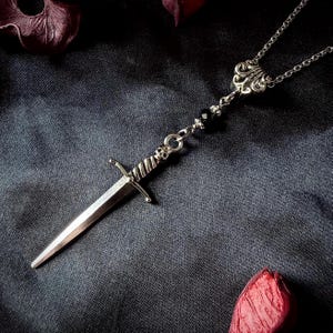Pode incluir: Um colar com pingente de adaga prateada. O pingente apresenta uma espada em miniatura detalhada com uma empunhadura em forma de cruz e uma lâmina pontiaguda. O colar tem uma corrente delicada com detalhes de contas pretas e um fecho ornamentado.