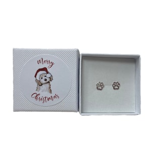 Op de afbeelding: Een witte geschenkdoos met een 'Merry Christmas' sticker met een hond met een kerstmuts. Binnenin zijn twee zilveren oorringen in de vorm van een pootafdruk te zien. De doos is open.