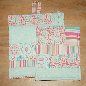 Puede incluir: Dos agarraderas de olla acolchadas con un diseño de patchwork en tonos de rosa, turquesa y blanco. La agarradera más grande tiene un lazo para colgar.