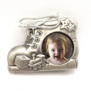 May include: Silver-toned baby boot photo frame ornament with a picture of a baby in the centre. The frame has a silver cord for hanging and features a toy train and block design.