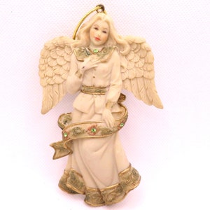 May include: A cream-colored angel ornament with gold wings and a green ribbon. The angel is wearing a white dress with gold trim and has green gem accents.