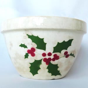 May include: A cream-colored ceramic mixing bowl featuring a decorative holly pattern with dark green leaves and clusters of bright red berries. The bowl has a wide, thick rim and a smooth, rounded body with a subtle, weathered, or aged finish.