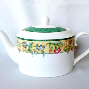 May include: A white ceramic teapot featuring a cylindrical body and a matching lid with a knob. A decorative band encircles the upper portion, displaying a yellow background with colourful floral illustrations in shades of pink, blue, and green.