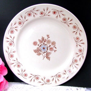 May include: A white ceramic dinner plate with a brown floral design. The plate has a scalloped edge and a raised rim.