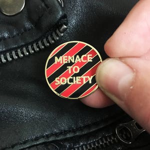 May include: A gold-colored pin with red and black stripes. The pin says "MENACE TO SOCIETY" in black letters.