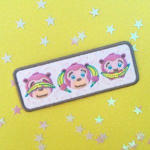 Könnte beinhalten: Ein gestickter Aufnäher mit drei rosa Comic-Affen, die gelbe Bananenschalen tragen. Die Affen lächeln und haben unterschiedliche Gesichtsausdrücke. Der Aufnäher ist auf weißem Hintergrund mit grauem Rand.