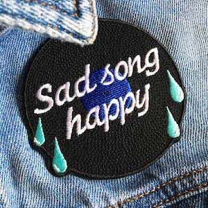 Könnte beinhalten: Schwarzer gestickter Bügeleisen-Aufnäher in Form einer Schallplatte mit dem Text "Sad song happy" in weißer und blauer Schrift und drei türkisfarbenen Tropfenformen.