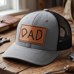 May include: Gray trucker hat with a black mesh back and a brown leather patch that reads "DAD" in black letters. The hat is sitting on a wooden surface with tools in the background.
