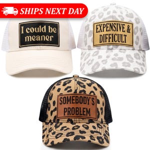 May include: Three trucker hats with different designs. One is cream-colored with the text "I could be meaner". Another is white with a leopard print and the text "Expensive & Difficult". The third hat has a leopard print and the text "Somebody's Problem".