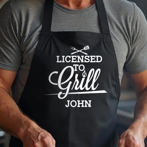 May include: Black apron with white text that reads "Licensed to Grill" with a flame graphic and the name "John" underneath.