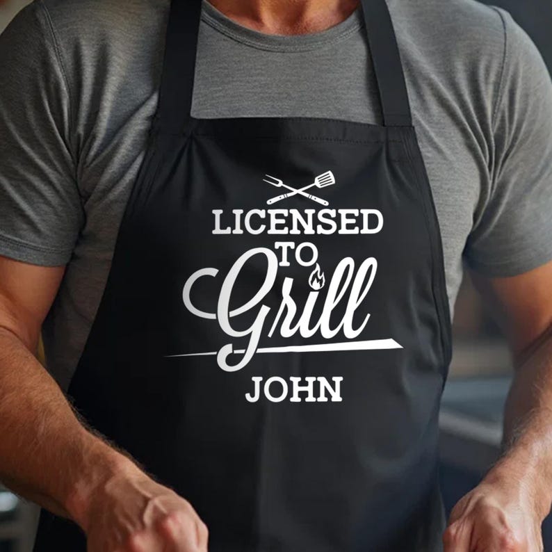 May include: Black apron with white text that reads "Licensed to Grill" with a flame graphic and the name "John" underneath.