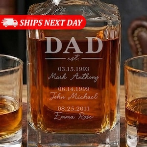 May include: A square glass decanter filled with amber liquid, with the word "DAD" etched on the front. Below the word "DAD" are the words "est." and three lines of text with names and dates. Two glasses of amber liquid are on either side of the decanter.