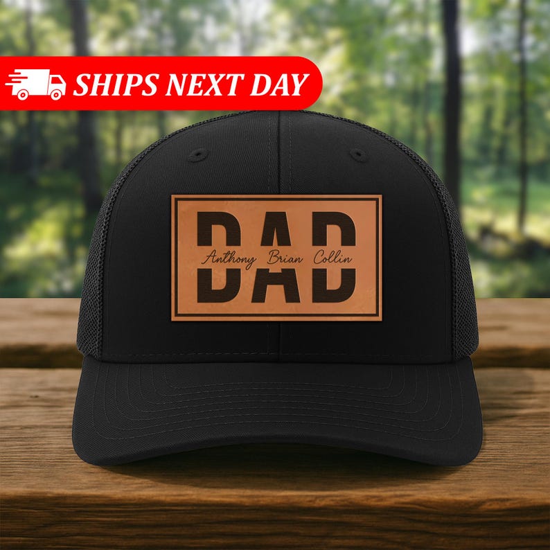 K&ouml;nnte beinhalten: Schwarze Trucker-Kappe mit Mesh-R&uuml;ckseite und gebogenem Schirm. Die Kappe hat einen rechteckigen Aufn&auml;her mit dem Wort "DAD" in fetten Buchstaben und dem Namen "Anthony Brian Collin" darunter. Der Aufn&auml;her ist hellbraun.