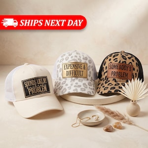 May include: Three trucker hats with mesh backs. The left hat is cream with a black patch reading "SOUNDS LIKE MY HUSBANDS PROBLEM". The center hat is grey leopard print with a gold patch reading "EXPENSIVE & DIFFICULT". The right hat is brown leopard print with a brown patch reading "SOMEBODY'S PROBLEM".