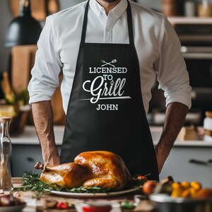 May include: Black apron with white text that reads "Licensed to Grill John". A person is wearing the apron and standing in a kitchen.