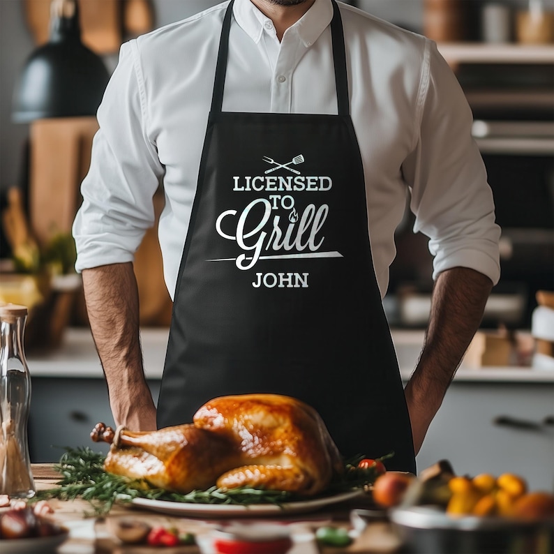 May include: Black apron with white text that reads "Licensed to Grill John". A person is wearing the apron and standing in a kitchen.