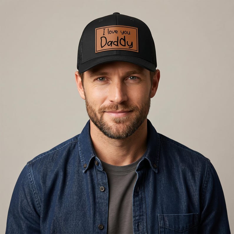 May include: Black trucker hat with a brown leather patch that reads "I love you Daddy" in black script. The hat has a mesh back and a curved brim. The hat is worn by a person.