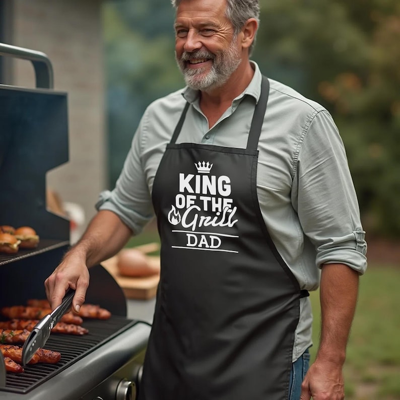 May include: A black apron with white text that reads "King of the Grill Dad" with a crown above the word "King".