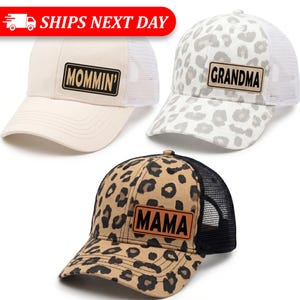 May include: Three baseball caps with mesh backs and text patches. One is beige with "Mommin'" on a black patch. Another is white with gray leopard print and "Grandma" on a black patch. The third is leopard print with "Mama" on a black patch.