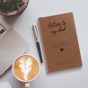 May include: A brown leather journal with the text "letters to my dad" and "all the things I wish I could tell you" in black lettering. A small heart symbol is below the text. The text "love, Meredith" is at the bottom of the journal.