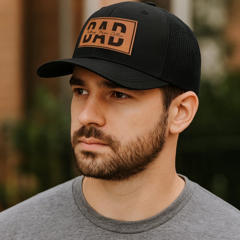 K&ouml;nnte beinhalten: Schwarze Baseballkappe mit Mesh-R&uuml;ckseite und gebogenem Schirm. Die Kappe hat einen braunen Lederaufn&auml;her mit dem Wort "DAD" in fetten, schwarzen Buchstaben. Darunter steht der Text "Anthony Brian Collin".