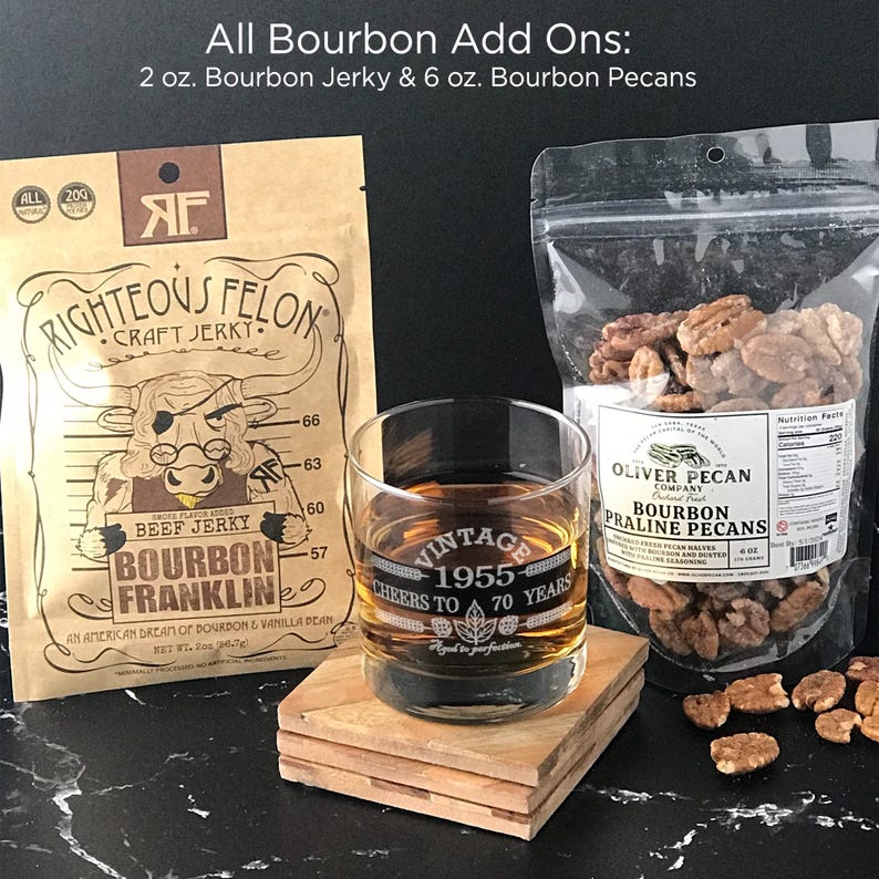 May include: A gift set featuring bourbon-flavored jerky and pecans. The jerky is packaged in a brown paper bag with the text "Righteous Felon Craft Jerky" and "Bourbon Franklin". The pecans are packaged in a clear plastic bag with the text "Oliver Pecan Company Bourbon Praline Pecans". The set also includes a glass with the text "Vintage 1955 Cheers to 70 Years".