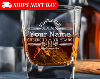 Puede incluir: Un vaso de whisky transparente lleno de líquido ámbar y cubitos de hielo. El vaso está grabado con el texto VINTAGE XXXX, Your Name, CHEERS TO XX YEARS y Aged to perfection, con gráficos decorativos de lúpulo y hojas en la parte frontal.