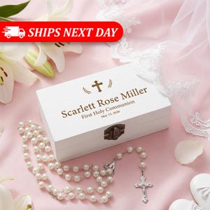 May include: A white wooden keepsake box with the text "Scarlett Rose Miller First Holy Communion May 15, 2026" and a cross design. A pearl rosary and a cross pendant are next to the box. The background is pink with white flowers.