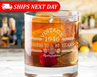 Puede incluir: Un vaso transparente lleno de un líquido color ámbar, cubitos de hielo y una cereza roja brillante. El vaso presenta un texto grabado en blanco que dice VINTAGE 1946 CHEERS TO 80 YEARS, acompañado de un diseño gráfico de una hoja de arce y lúpulo.