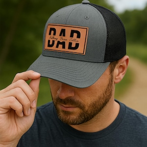 K&ouml;nnte beinhalten: Graue Trucker-Kappe mit schwarzem Mesh-R&uuml;cken. Die Kappe hat einen braunen Lederaufn&auml;her mit dem Wort "DAD" in fetten, schwarzen Buchstaben und den Namen "Anthony Brian Collin" darunter.