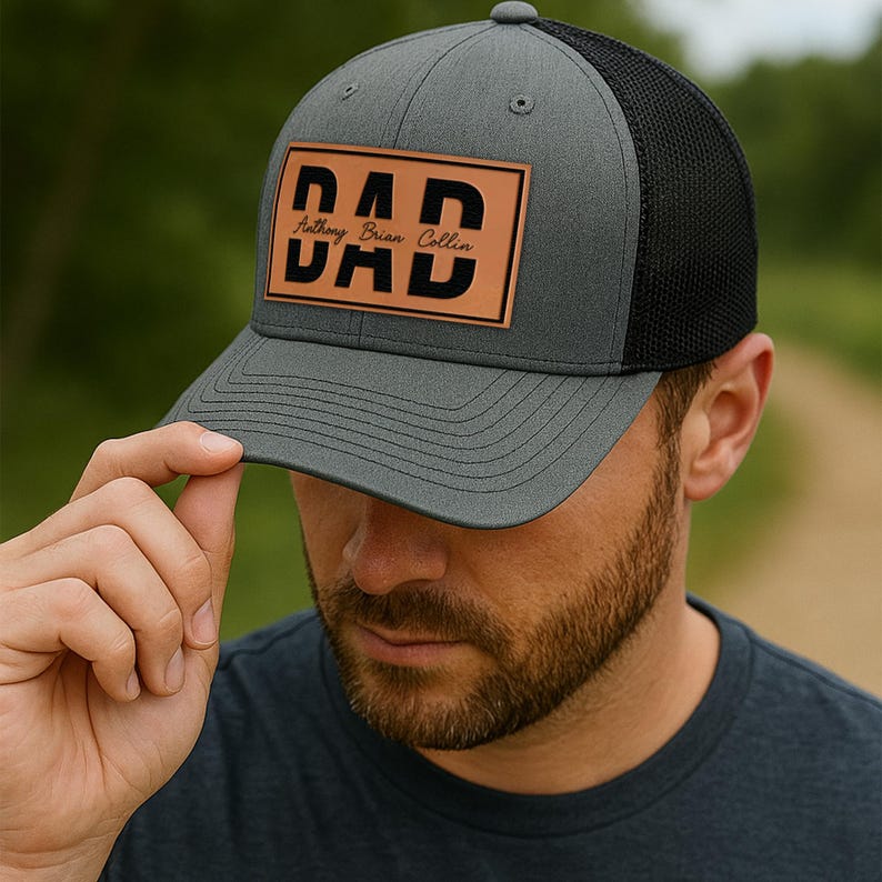 K&ouml;nnte beinhalten: Graue Trucker-Kappe mit schwarzem Mesh-R&uuml;cken. Die Kappe hat einen braunen Lederaufn&auml;her mit dem Wort "DAD" in fetten, schwarzen Buchstaben und den Namen "Anthony Brian Collin" darunter.