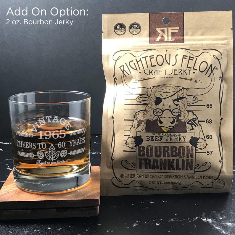 May include: A brown paper bag with a cartoon bull wearing a bandana and glasses. The bag is labeled "Righteous Felon Craft Jerky" and features the text "Bourbon Franklin" and "Smoke Flavor Added Beef Jerky". The bag is sitting on a wooden coaster next to a clear glass with a brown liquid inside. The glass has the text "Vintage 1965 Cheers to 60 Years" etched on it.