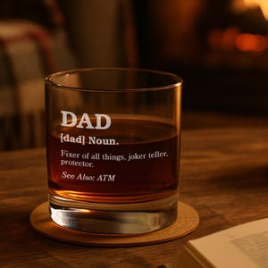 Puede incluir: Un vaso de vidrio transparente con la palabra "DAD" grabada en la parte delantera, junto con una definici&oacute;n. El vaso est&aacute; lleno de un l&iacute;quido marr&oacute;n y descansa sobre un posavasos de madera. El texto dice "Arreglador de todo, cuentachistes, protector."
