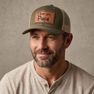 May include: Olive green and tan trucker hat with the text "I love you Papa" on a brown leather patch. The hat has a curved brim and a mesh back.