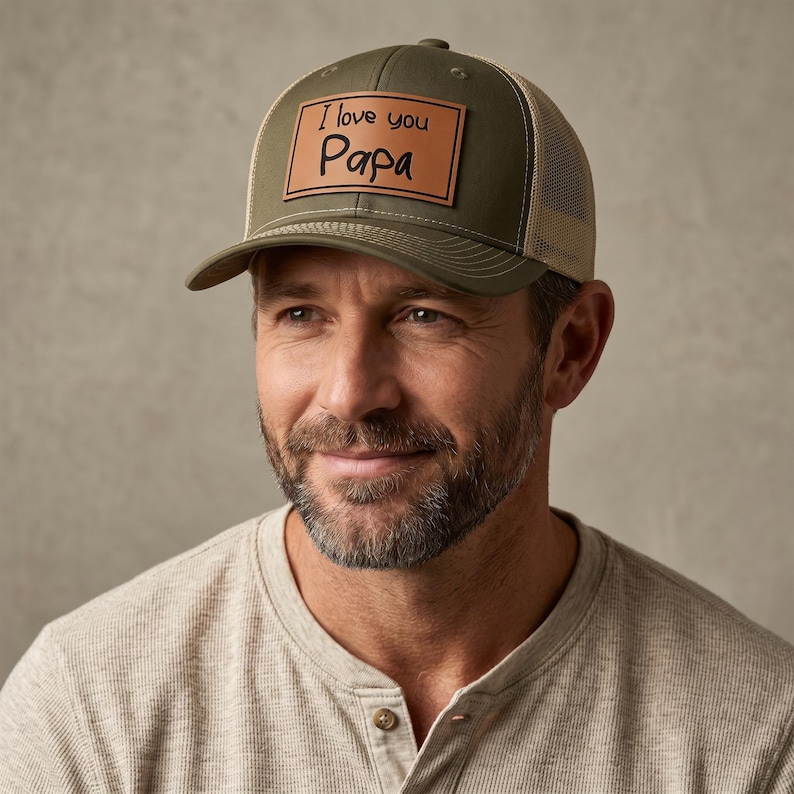 May include: Olive green and tan trucker hat with the text "I love you Papa" on a brown leather patch. The hat has a curved brim and a mesh back.