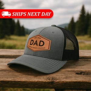 May include: Gray and black trucker hat with a leather patch that reads "DAD" in bold letters. The patch also includes the text "EST. 1995" and names. The hat is sitting on a wooden surface.