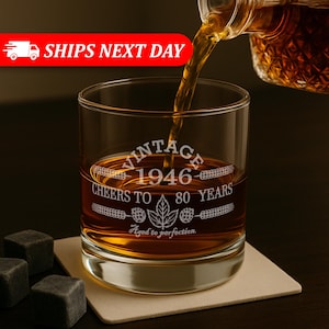 May include: A clear glass tumbler filled with amber liquid being poured from a bottle. The glass features white etched text reading VINTAGE 1946 CHEERS TO 80 YEARS with a maple leaf and wheat graphic, plus the phrase Aged to perfection.