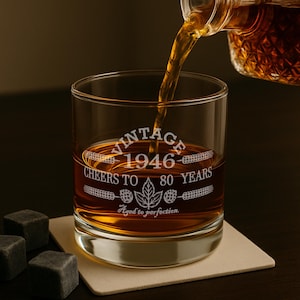May include: A clear glass tumbler filled with amber liquid being poured from a bottle. The glass features white etched text reading VINTAGE 1946 CHEERS TO 80 YEARS with a leaf and hop graphic and the phrase Aged to perfection below the design.