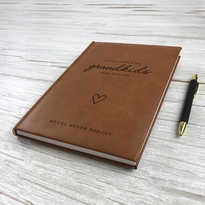 K&ouml;nnte beinhalten: Ein braunes Notizbuch mit texturierter Oberfl&auml;che, bedruckt mit dem schwarzen Text CUTE THINGS MY grandkids SAY AND DO, einem kleinen Herzsymbol und dem Text SO I'LL NEVER FORGET. Ein schwarzer Stift mit goldenen Akzenten liegt rechts daneben.