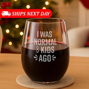 May include: A stemless wine glass filled with red wine, resting on a beige coaster. The glass has the text "I WAS NORMAL 3 KIDS AGO" in white. The background includes a Christmas tree with lights.