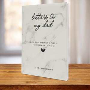 May include: A white marble-patterned journal with the text "letters to my dad" and "ALL THE THINGS I WISH I COULD TELL YOU" in black. A small black heart is below the text. The bottom of the journal says "LOVE, MEREDITH".