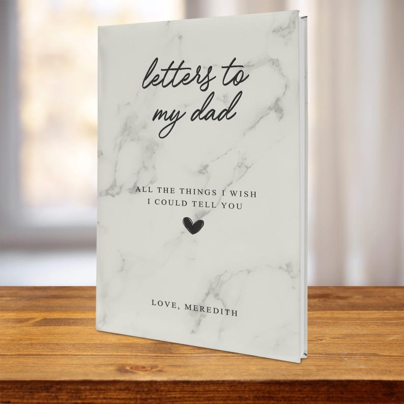May include: A white marble-patterned journal with the text "letters to my dad" and "ALL THE THINGS I WISH I COULD TELL YOU" in black. A small black heart is below the text. The bottom of the journal says "LOVE, MEREDITH".