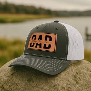 K&ouml;nnte beinhalten: Grau-wei&szlig;e Trucker-Cap mit einem Lederaufn&auml;her mit dem Wort "DAD" in gro&szlig;en schwarzen Buchstaben. Der Aufn&auml;her enth&auml;lt auch den Namen "Anthony Brian Collin". Die Kappe liegt auf einem Felsen.