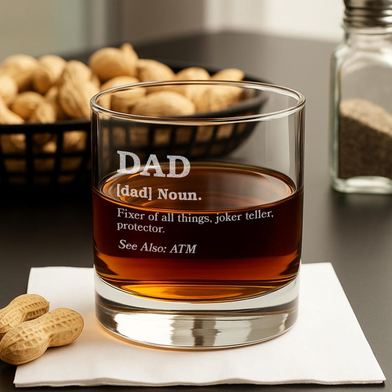 Puede incluir: Un vaso transparente lleno de l&iacute;quido &aacute;mbar, con la palabra "DAD" grabada. La definici&oacute;n de "DAD" tambi&eacute;n est&aacute; grabada en el vaso. El vaso est&aacute; sobre una servilleta blanca, con cacahuetes y un salero en el fondo.